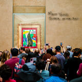 A photograph of a group of people in an art museum crowded around a child's drawing which is behind glass.